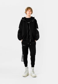 Joven de pie vestido con sudadera con capucha negra y pantalones deportivos con parches gráficos blancos, combinados con botas blancas con cordones, sobre un fondo liso.