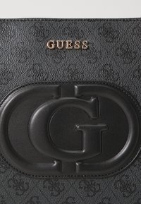 Borsa nera in pelle sintetica con texture, dotata di un logo circolare "G" in rilievo e un logo metallici "GUESS" in rosa oro nella parte superiore.