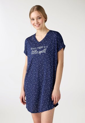 Giovane donna sorridente, indossa una camicia da notte blu navy con costellazioni di stelle bianche e testo: "Ogni notte è un piccolo incantesimo."