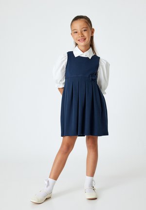Marineblaues Faltenkleid mit Schleifendetail, weißen Puffärmeln und einem Kragenhemd. Kombiniert mit weißen Socken und hellen Schuhen.