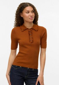 Femme aux cheveux bouclés portant un pull à manches courtes en maille côtelée de couleur rouille, avec un col volé et une patte de boutonnage, associé à un jean bleu foncé.
