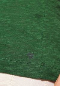T-shirt corto verde scuro realizzato in un morbido tessuto testurizzato. Presenta un piccolo logo viola vicino all'orlo. Cuciture piatte senza etichette visibili.