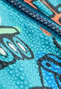 Tkanina odporna na wodę w kolorze turkusowym z kroplami wody. Widoczny czarno-pomarańczowy akcent graficzny. Pokazane detale zamka.