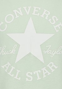 Világos zöld pamut póló, amelyen egy nagy fehér csillag grafika látható a "CONVERSE" és az "ALL STAR" felirattal, kiemelve a letisztult, merész tipográfiát.