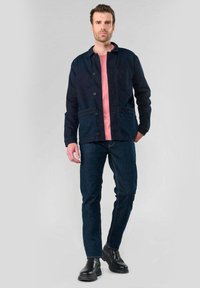 Marineblauwe button-up jas met twee voorzakken, gelaagd over een roze shirt, gecombineerd met donkerblauwe jeans en zwarte enkellaarsjes.
