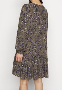 Ruskea mekko, jossa on pitkät kerrottu hihat, leopardikuvio purppurassa ja sinisessä, röyhelöhelma ja väljä istuvuus, esittelee pehmeää kangasrakennetta.