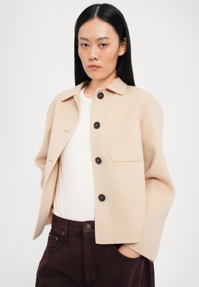Chaqueta de forro polar beige con un cuello, botones negros y un único bolsillo en el pecho; llevada sobre una camiseta blanca y combinada con pantalones burdeos.