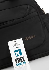 Černá nylonová taška s kapsou na zip, vybavená kovovou značkovou plaketou a štítkem s nápisem "FREE CARRY-ON". Hladká textura, minimalistický design.