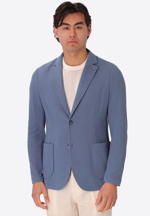 Uomo che indossa un blazer azzurro chiaro strutturato sopra una camicia bianca e pantaloni chiari, in piedi davanti a uno sfondo bianco uniforme.