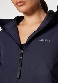 Granatowa kurtka z kapturem, zapinana na zamek, z czarnymi elementami. Posiada gładką teksturę, logo na piersi oraz regulowany kaptur ze sznurkiem.