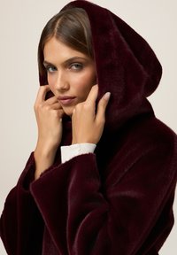 Cappotto cappuccio borgogna in morbido tessuto peluche con un ampio colletto e vestibilità rilassata. La superficie texturizzata aggiunge profondità, completata da dettagli bianchi sui polsini.
