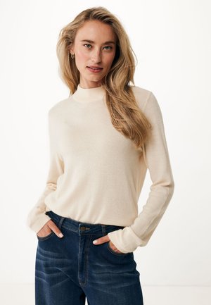 Suéter beige de manga larga con cuello alto, de textura ligera y puños ajustados, combinado con unos jeans azul oscuro que tienen bolsillos delanteros.