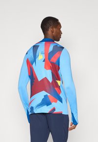 Koszulka sportowa z długim rękawem w jasnoniebieskim kolorze z abstrakcyjnym wzorem z czerwonymi, granatowymi i żółtymi akcentami; gładka faktura i dopasowany krój.