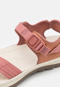 Vaaleanpunainen sandaali, jossa on teksturoitu hihna, säädettävä solki ja vaaleanvärinen anatominen jalkapohja. Kumipohja tarjoaa pidon.