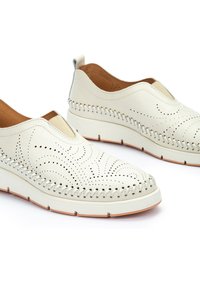 Zapatos tipo slip-on de cuero blanco con diseños perforados, detalles de costura en contraste alrededor de la suela y un interior texturizado en tono marrón.