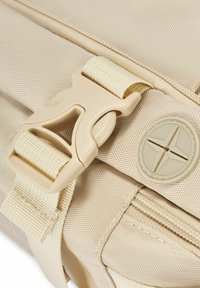 Szczegół beżowej torby z solidnym klipsem i teksturowanym materiałem, z okrągłym emblematem i kontrastowym wzorem paska dla funkcjonalności.