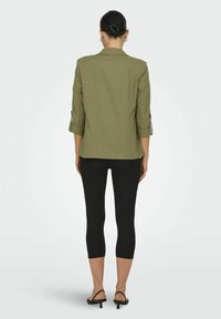 Blazer verde oliva con maniche arrotolate, chiusura a un bottone e vestibilità strutturata, abbinato a leggings neri e sandali con tacco basso.