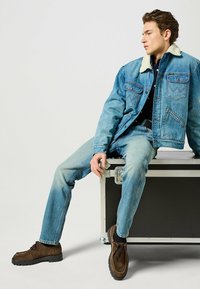 Giacca di jeans con colletto in pelliccia sintetica, lavaggio azzurro chiaro sbiadito; jeans abbinati; scarpe in camoscio marrone con suole nere spesse; seduto su una cassa.