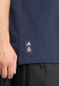 Marineblaues Baumwoll-T-Shirt mit einem Adidas-Logo und dem Real Madrid Wappen auf einem rechteckigen Abzeichen am Saum. Schwarze Shorts teilweise sichtbar.