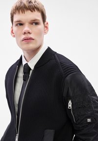 Casaco bomber preto com textura canelada, fecho de correr e mangas com acabamento acetinado com bolso de fecho. Camisa branca e gravata visíveis por baixo.