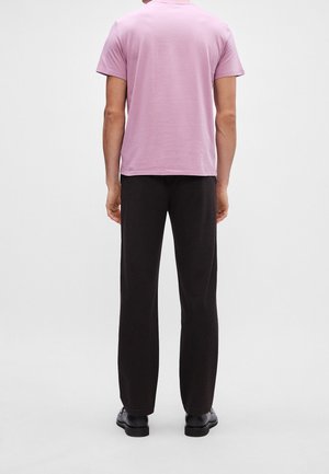 Mand iført en pink t-shirt med korte ærmer, sorte bukser og sorte sko, stående med ryggen til kameraet på en ensfarvet baggrund.