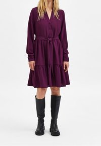 Lila skjortklänning med långa ärmar, midjeband och flerlagerskjol. Bärs med svarta knähöga stövlar, slät textur och flödande design.