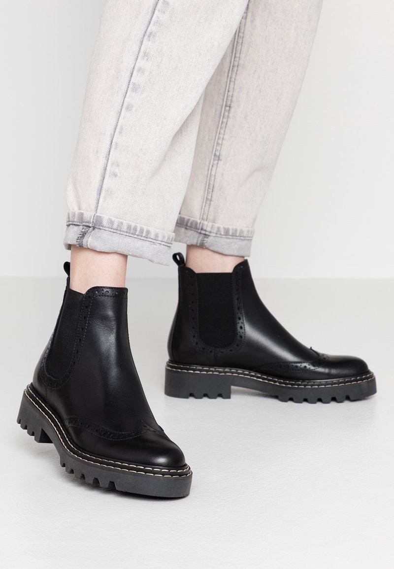 Bottines en cuir noir avec panneaux élastiques sur les côtés, broguing décoratif, semelle en caoutchouc épaisse et coutures contrastantes.