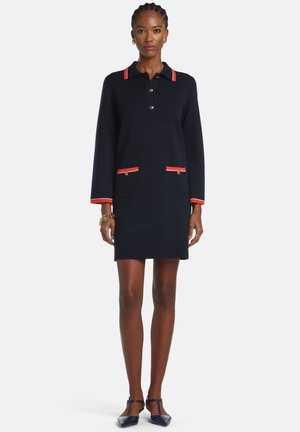 Kobieta ubrana w granatową sukienkę polo z długimi rękawami, z czerwonym wykończeniem na kołnierzyku, mankietach i kieszeniach, stojąca z rękami swobodnie opuszczonymi wzdłuż ciała.