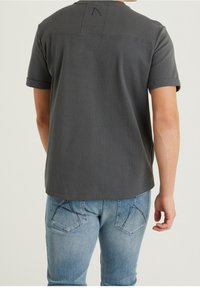 Graues Mesh-Shirt mit kurzen Ärmeln und umgeschlagenen Ärmeln, mit strukturiertem Finish und dezentem Logo-Detail auf der Rückseite. Getragen mit blauen Jeans.