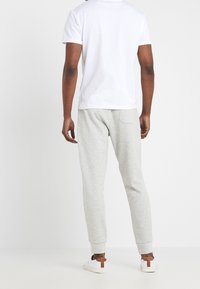 Miesten valkoinen t-paita, vaaleanharmaat collegehousut joustavilla resoreilla, sileää kangasta ja takataskun yksityiskohta. Valkoiset tennarit ruskein yksityiskohdin.