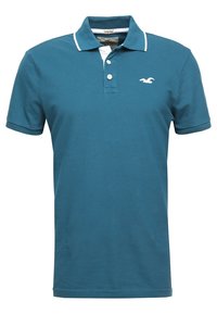 Teal-poolopaita joustavaa kangasta, jossa on klassinen kaulus, kaksi nappia ja valkoinen logo-aksentti vasemmassa rinnassa.