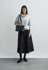 Grå polotröja, svart knälång kjol med texturerade blommönster, svarta klackskor och en strukturerad mörk väska med spänne.