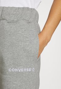 Šedé tepláky s elastickým pasem a bočními kapsami, zdobené bílou výšivkou loga "CONVERSE" poblíž kapsy. Měkká textilie.