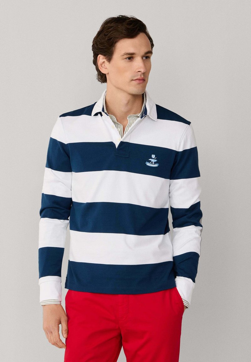 Uomo che indossa una polo a maniche lunghe con righe orizzontali blu navy e bianche, pantaloni rossi e una camicia con colletto sotto, con le mani in tasca, che guarda verso destra.