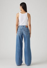 Pantalones vaqueros de tiro alto en denim azul de pierna ancha, con un parche de cuero en la parte posterior y dos bolsillos de parche, mostrados desde atrás.