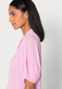 Blusa rosa com mangas curtas e volumosas, apresentando um decote com botões e um padrão de bolinhas brancas, fabricada em tecido leve e texturizado.