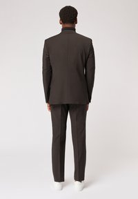 Dunkelbrauner Anzug mit taillierter Passform, hohem Kragen und schmalen Hosen, kombiniert mit weißen Sneakers. Glatte Textur, minimale Akzente.