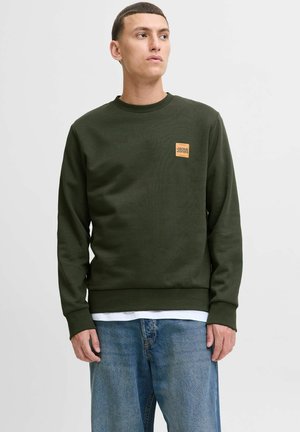 Un jeune homme portant un sweat à capuche vert olive avec un petit logo orange Jack & Jones sur la poitrine, associé à un jean bleu, sur fond blanc.