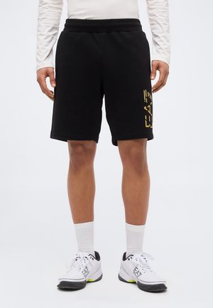 Mann trägt schwarze Shorts mit gelbem Motiv auf dem Oberschenkel, weißes Langarmshirt, weiße Kniestrümpfe und weiße Sportschuhe mit schwarzen Sohlen.