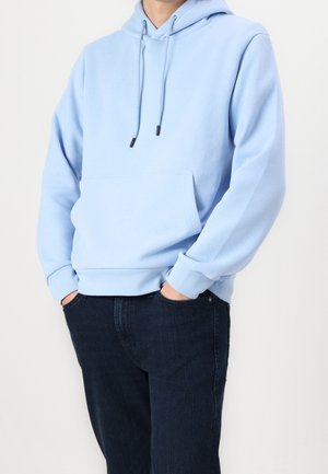 Homme portant un sweat à capuche bleu clair avec poche avant et cordons, associé à un jean bleu foncé sur fond blanc.