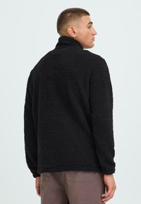 Chaqueta de forro polar negra con cuello alto, tela texturizada, mangas largas y corte holgado, mostrada desde un lado y una vista trasera.