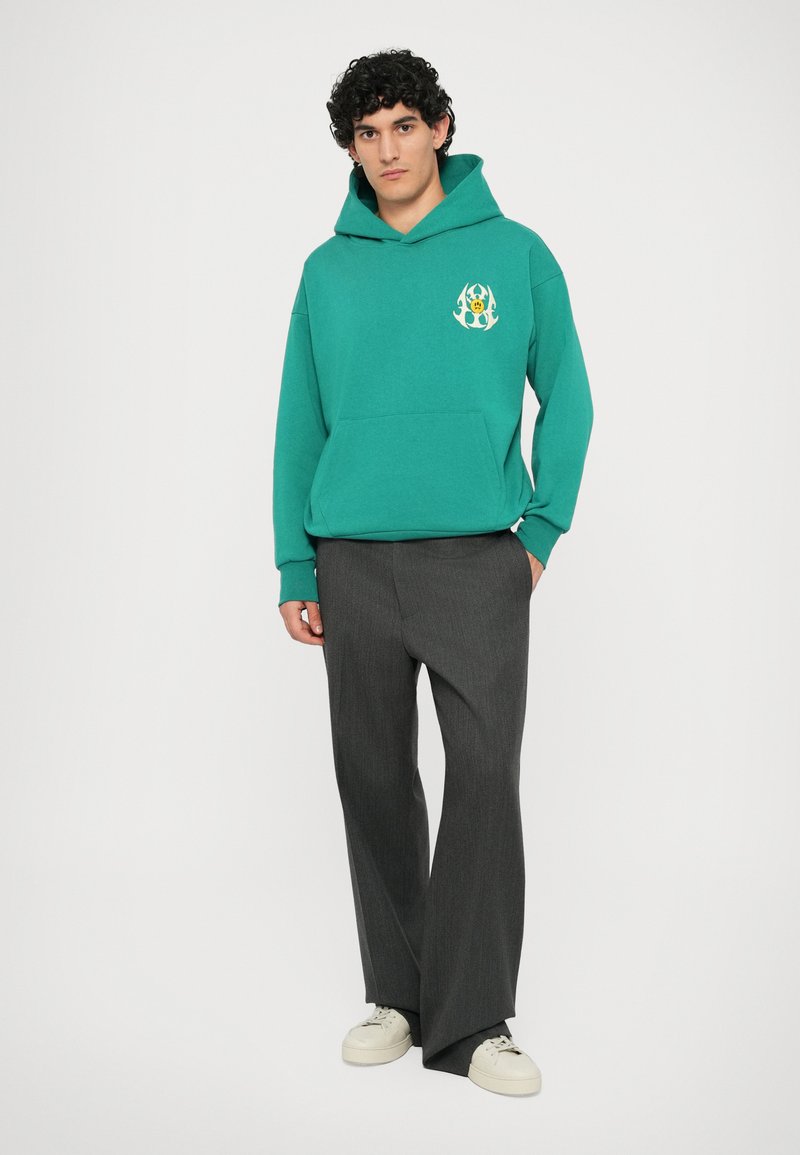 Felpa turchese con tasca frontale e logo, abbinata a pantaloni larghi grigi e sneakers bianche. Presenta un cappuccio con cordino.