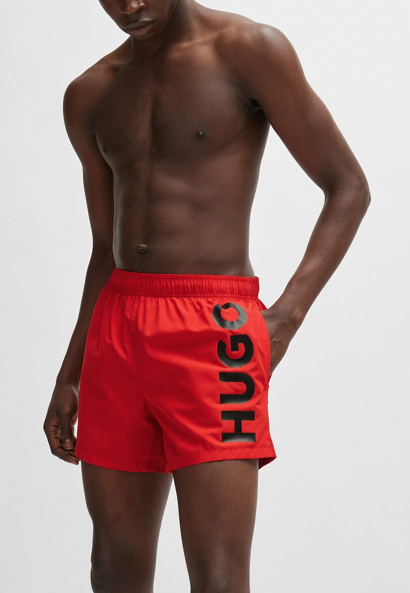Red swim shorts featuring an elastic waistband and side pockets, branded with bold black "HUGO" lettering along the left side.