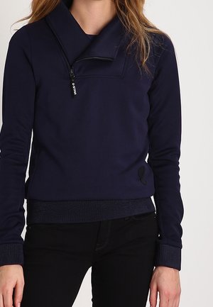 Pullover blu navy con colletto alto e tasca con zip. Realizzato in tessuto liscio con polsini e orlo a coste. Presenta un dettaglio logo laterale.