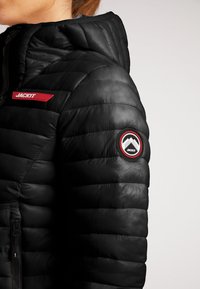 Giacca a vento nera con cappuccio, caratterizzata da un patch logo rosso e un emblema rotondo sulla manica. Chiusura con zip visibile sul davanti.