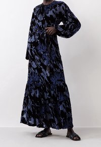 Robe en velours floral noire et bleue, longue, avec des manches longues, un col froncé et une taille cintrée. Jupe évasée avec un ourlet à volants. Sandales noires.