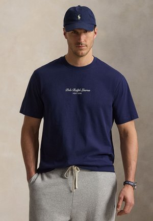 Homme portant une casquette Polo Ralph Lauren bleu marine et un t-shirt bleu marine assorti avec logo, un pantalon de survêtement gris clair avec cordon de serrage, et une montre.