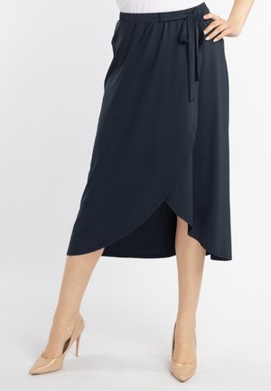 Femme portant une jupe midi portefeuille bleu marine avec un lien sur le côté et des chaussures à talons beiges, debout avec une main sur la hanche.