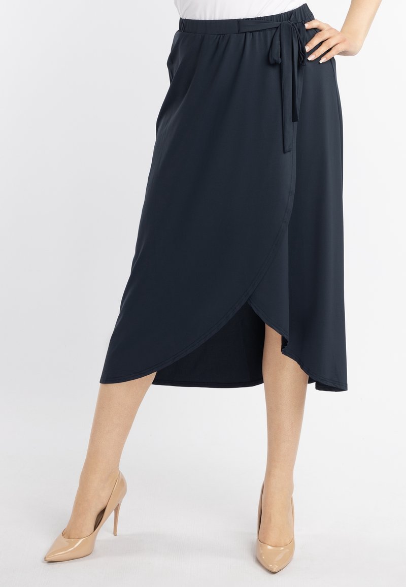 Femme portant une jupe midi portefeuille bleu marine avec un lien sur le côté et des chaussures à talons beiges, debout avec une main sur la hanche.
