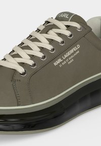 Baskets Karl Lagerfeld vert olive avec lacets blancs, œillets argentés, nom et adresse de la marque sur le côté, et une semelle noire transparente.